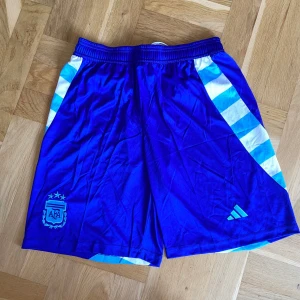 Blå Adidas Argentina fotbollsshorts - Snygga blå fotbollsshorts från Adidas med Argentina AFA-logga broderad på ena benet. De har randiga detaljer i vitt och ljusblått längs sidorna och elastisk midja med snörning. Perfekta för dig som älskar fotboll och vill ha en sportig look.