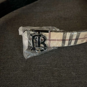 Beige rutigt Burberry bälte - Snyggt bälte från Burberry med klassiskt beige rutigt mönster och en stor silvrig B-spänne framtill. Bältet har en lyxig känsla och är gjort i skinn med detaljer i svart och brunt. Perfekt accessoar för att lyfta din outfit.