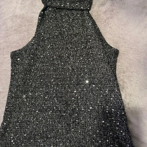 Glittrig svart ärmlös topp - Supersnygg svart ärmlös topp med glittriga detaljer. Perfekt för dig som vill sticka ut och glänsa på nästa fest. Hög hals och figurnära passform ger en trendig look. Materialet är stretchigt och bekvämt.