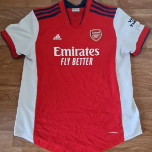 Arsenal tröja direkt från England i butik 2021 kit  - Snygg Arsenal fotbollströja från Adidas i storlek XL. Tröjan är röd med vita paneler på sidorna och ärmarna, samt blå detaljer på axlarna. Klassisk Arsenal-logga på bröstet och 'Emirates Fly Better' tryck på framsidan. Tillverkad i lätt och ventilerande polyester.