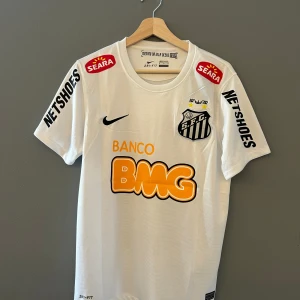 Vit fotbollströja från Nike - Säljer en vit fotbollströja från Nike med Santos FC-logga och sponsortryck i svart och orange. Tröjan har korta ärmar och Neymar Jr:s namn och nummer 11 på ryggen. Perfekt för fotbollsfans! Helt ny med tagg i storlek M en replika