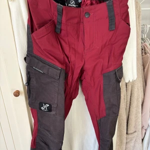 Röda och svarta byxor från RVRC - Säljer ett par funktionella GPX Pants från RVRC i röd och svart färg. Byxorna har flera praktiska fickor med dragkedja och knappar, samt förstärkta partier för extra hållbarhet. Perfekta för utomhusaktiviteter.
