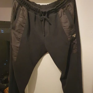 Svarta mjukisbyxor från Prada - Snygga svarta mjukisbyxor från Prada med elastisk midja och snörning. Byxorna har detaljer i form av vadderade paneler och en diskret logotyp på sidan. Perfekta för en avslappnad stil.Det ni ser på bilden är det ni får😁 ett plagg som är köpt i bulk så jag vet lila lite som ni, men jag har kollat så gott jag kan på alla möjliga sajter och det jag kan kolla stämmer överens med originalet väldigt bra tröja raka o fina sömmar, kvalitets lappar och tyg