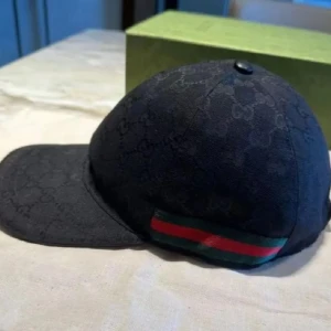 Svart Gucci keps - Snygg svart keps från Gucci med diskret mönster och klassiska gröna och röda ränder på sidan. Perfekt för att ge din outfit en lyxig touch.