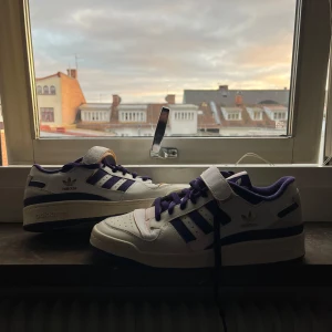 Adidas Forum low sneakers i vitt och lila - Snygga Adidas sneakers i vitt med lila detaljer och snörning. Skorna har en klassisk design med tre ränder på sidan och en kardborreband över snörningen för extra stil. Perfekta för en sportig look. Helt nya. Var använda några gånger 