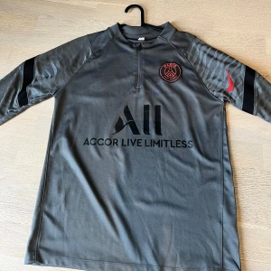 Psg kofta  - Snygg grå fotbollströja från Nike med PSG-logga på bröstet och 'Visit Rwanda' på ryggen. Tröjan har svarta och röda detaljer samt korta ärmar. Perfekt för fotbollsfans! Kan sänka pris vid snabb affär