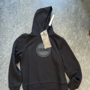 Svart hoodie från Colmar - Säljer en stilren svart hoodie från Colmar med deras ikoniska logga på bröstet. Tröjan har en bekväm passform med långa ärmar och en praktisk huva. Perfekt för en avslappnad look. Står storlek L men passar storlek M bättre.