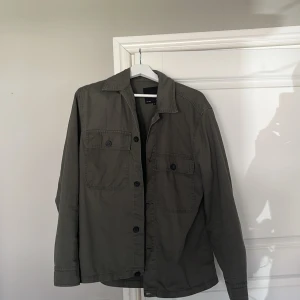 Grön overshirt från 157 - Säljer en stilren grön overshirt från 157. Jackan har långa ärmar och knappar framtill samt två bröstfickor med knappar. Perfekt för lager-på-lager-stil.