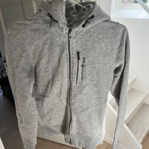 Grå hoodie från Sail Racing - Säljer en stilren grå hoodie från Sail Racing med dragkedja och justerbar huva. Tröjan har en praktisk bröstficka med dragkedja och ribbade muddar vid ärmslut och nederkant. Perfekt för en avslappnad look.