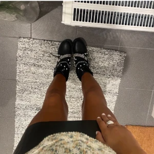 Svarta boots med kedjedetaljer - Snygga svarta boots med kedjedetaljer och spännen. Skorna har snörning och är dekorerade med nitar för en tuff look. Perfekta för att ge din outfit en edgy touch.