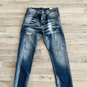 Dsquared2 jeans - Snygga blå jeans med coola slitningar och färgstänk. De har en normal passform och är perfekta för en avslappnad stil. Jeansen har en klassisk femficksdesign och en knappgylf. Storlek 48