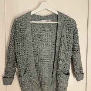 Grön stickad kofta från 157 - Säljer en mysig grön stickad kofta från 157. Koftan har ett öppet mönster och två praktiska fickor framtill. Perfekt för kyliga dagar och ger en avslappnad look. Passar bra till både jeans och klänning.