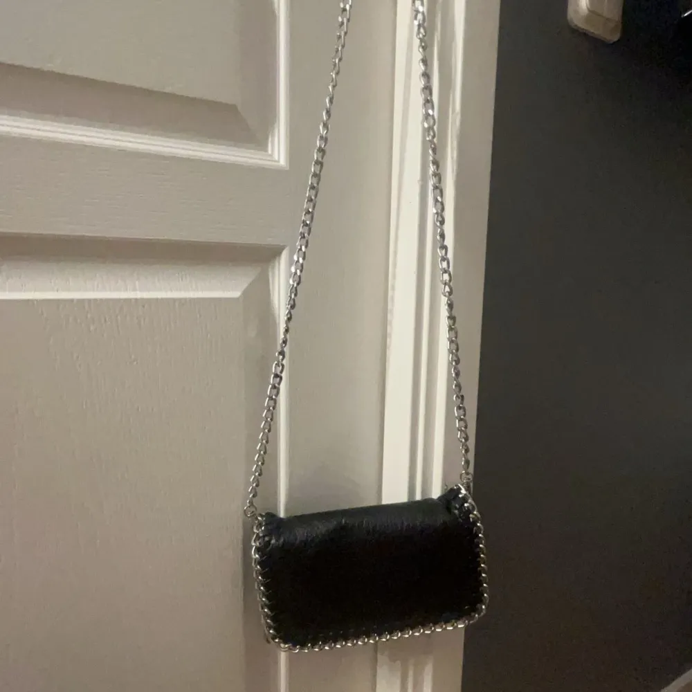 Snygg svart axelväska från Tiamo i den minsta modellen med kedjerem och dekorativa kedjedetaljer längs kanterna. Perfekt för att ge en stilren touch till din outfit. Köpt på scorett för 500kr. Laukut & Käsilaukut.