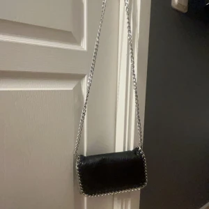 Svart axelväska från Tiamo - Snygg svart axelväska från Tiamo i den minsta modellen med kedjerem och dekorativa kedjedetaljer längs kanterna. Perfekt för att ge en stilren touch till din outfit. Köpt på scorett för 500kr