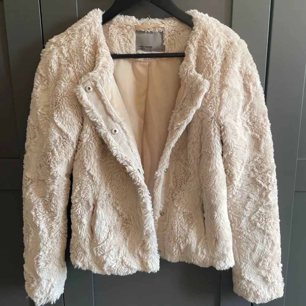 Beige pälsjacka i fuskpäls från Vero Moda. Knappar framtill och en ficka per sida. Kort modell.. Takit.