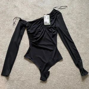 Svart oneshoulder bodysuit - Snygg svart oneshoulder bodysuit med långa ärmar och rynkdetaljer. Perfekt för en stilren look. Tillverkad i ett mjukt och stretchigt material för bästa komfort.