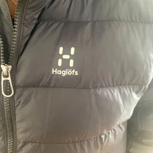Svart dunjacka från Haglöfs - Säljer en svart dunjacka från Haglöfs med dragkedja och huva. Jackan är perfekt för kalla dagar och har en stilren design med Haglöfs logga på bröstet.