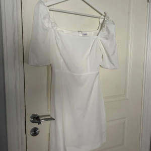 Vit klänning med puffärmar - Säljer en stilren vit klänning med puffiga kortärmar och en elegant passform. Perfekt för sommarens alla tillfällen. Klänningen har en fyrkantig urringning och är gjord i ett lätt material. Använd 1 gång!