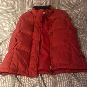 Röd dunväst med dragkedja - Snygg röd dunväst med dragkedja och praktiska fickor. Perfekt för kyliga dagar och ger en sportig look. Västen har en hög krage för extra värme.
