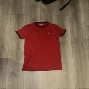 Röd t-shirt från Lyle & Scott - Säljer en snygg röd t-shirt från Lyle & Scott med korta ärmar och en liten broderad logga på bröstet. T-shirten har en mörkare kant vid halsen som ger en fin kontrast. Perfekt för en avslappnad stil!