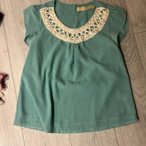 Grön blus med spetsdetaljer från Mina uk - Säljer en söt grön blus från Minku med vackra spetsdetaljer i beige runt halsen. Blusen har korta ärmar och en luftig passform, perfekt för varmare dagar. Den är tillverkad i ett lätt och bekvämt material.