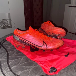 Röda Nike fotbollsskor - Säljer ett par röda Nike fotbollsskor med svarta detaljer och snörning. Skorna har en modern design med en tydlig Nike-logga på sidan. Perfekta för fotbollsplanen!