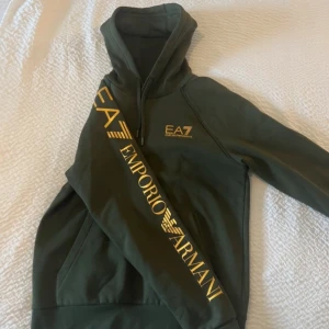 Grön hoodie från Emporio Armani - Snygg grön hoodie från Emporio Armani med gul logga på bröstet och ärmen. Den har en klassisk känguruficka och en bekväm huva med dragsko. Perfekt för en stilren och avslappnad look.   Storlek S-M