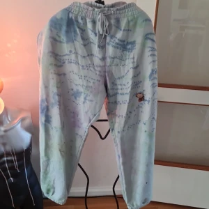 Mönstrade mjukisbyxor från New Balance - Säljer ett par bekväma mjukisbyxor från New Balance i en cool batikstil med blå och lila nyanser. Byxorna har en elastisk midja med snörning och ett broderat New Balance-märke på benet. Perfekta för en avslappnad stil!