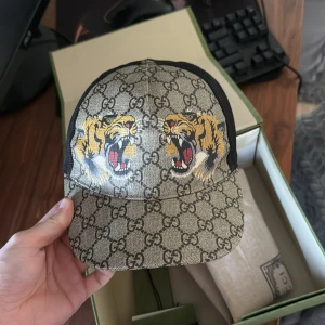 Gucci keps tiger, storlek S - Gucci keps med tiger tryck, köpt 2021 på farfetch, kepsen är i bra skick och endast använd ett par gånger. Medföljer dustbag, alla lappar och kuvert som man får när man köper den och box, säljer den pga att jag inte använder den längre. Priset är förhandlingsbart och inte fast i sten så kom med erat bästa pris.
