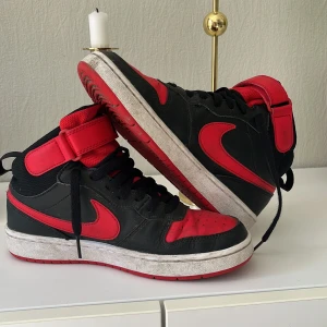 Svarta och röda Nike sneakers - Snygga svarta och röda Nike sneakers med hög design och klassisk snörning. Skorna har en Nike-logga på sidan och baksidan. Perfekta för en sportig look.