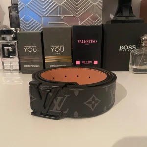 Svart bälte från Louis Vuitton - Snyggt svart bälte från Louis Vuitton med det ikoniska monogrammönstret i grått. Bältet har en stilren design med en svart spänne som pryds av LV-logotypen. Perfekt för att ge en lyxig touch till din outfit.