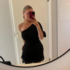 Svart glittrig klänning - Snygg svart klänning med en axel och glittrigt mönster. Perfekt för en kväll ute. Den har en volangdetalj vid fållen som ger en extra touch.