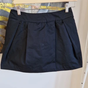 Svart kortkjol med tyll - Snygg svart kortkjol med ett lager av tyll under för extra volym. Kjolen har en klassisk design med veck framtill och en bekväm passform. Perfekt för att ge en stilren look med en twist.