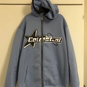 Blå hoodie med Celestial-tryck - Snygg ljusblå hoodie med dragkedja och stor svartvit Celestial-logga framtill. Tröjan har huva, känguruficka och långa ärmar. Perfekt för dig som gillar streetwear och vill sticka ut med ett coolt tryck. Helt ny, aldrig andvänd. Den är i flis material från insidan. 