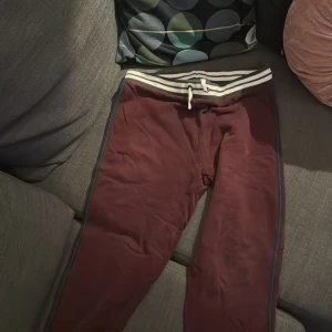 Vinröda mjukisbyxor med snörning - Snygga vinröda mjukisbyxor med vita och svarta ränder i midjan och snörning. Byxorna har en relaxed fit och blå sidodetaljer längs benen. Perfekta för chill dagar eller när du vill vara bekväm men ändå se fräsch ut.