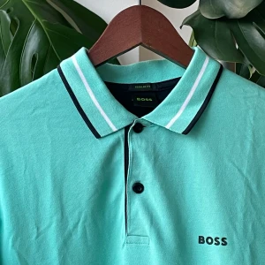 BOSS pikétröja - Storlek:S. Snygg turkos pikétröja från BOSS med svart och vit rand på kragen och ärmslut. Klassisk piké med två knappar och broderad BOSS-logga på bröstet. Perfekt för en fräsch och stilren look. Tillverkad i mjuk bomull för maximal komfort.