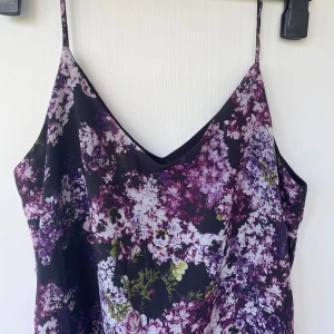 Blommig sommarklänning från Gina Tricot - Supersnygg klänning med smala axelband från Gina Tricot. Klänningen har ett lila, vitt och grönt blommönster på svart botten och en v-ringad halsringning. Perfekt för sommarens alla tillfällen!