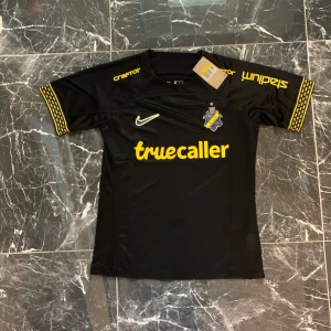 AIK svart matchtröja Nike 2024 - AIK:s officiella svarta matchtröja från Nike med gula detaljer och tryck. Tröjan har AIK:s klubbmärke på bröstet, sponsorer i gult och unika mönster på ärmsluten. Tillverkad i lätt och ventilerande funktionsmaterial, perfekt för fotbollsträning eller match.