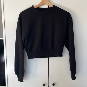 Svart croppad sweatshirt från H&M Divided - Svart croppad sweatshirt från H&M Divided med rund halsringning och breda muddar vid ärmslut och nederkant. Tröjan har en loose passform och är tillverkad i mjuk bomullsmix, perfekt för en chill och avslappnad stil.