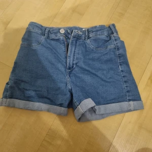 Blå jeansshorts med upprullad kant - Snygga blå jeansshorts med upprullad kant och klassisk femficksdesign. De har normal passform och stängs med dragkedja och knapp. Perfekta för varma dagar och enkla att matcha med allt. Tidlös modell som alltid funkar.