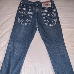 True Religion blå jeans med kontrastsömmar - Säljer ett par klassiska blå jeans från True Religion med tydliga vita kontrastsömmar och ikoniska bakfickor med lock och metallknappar. Jeansen är raka i modellen och tillverkade i 100% bomull. Perfekta för dig som gillar streetwear och vill sticka ut.