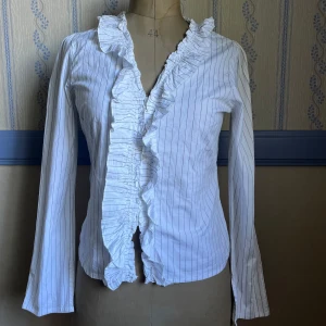 Vit randig blus med volangkrage - Jättesnygg vit skjorta med tunna svarta ränder och volang längs v-ringningen. Långärmad och bra passform. Passar perfekt för vardag och finare tillfällen. Är i storlek XL men tycker själv att den är som en L/M