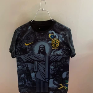 Svart Brasil fotbollströja Nike - Unik svart Brasilien fotbollströja från Nike med detaljerat grafiskt tryck av Kristusstatyn och brasilianska symboler. Gula loggor och emblem på bröstet, kortärmad modell i lätt och ventilerande material. Perfekt för dig som vill sticka ut på planen eller läktaren.