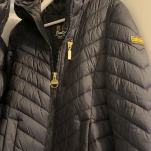 Svart Barbour pufferjacka med huva - Säljer en svart pufferjacka från Barbour International med snygga gulddetaljer på dragkedjan och logga på ärmen. Jackan har huva, quiltad design och dragkedja framtill. Perfekt för kalla dagar och riktigt stilren look. Har knappt använt den 