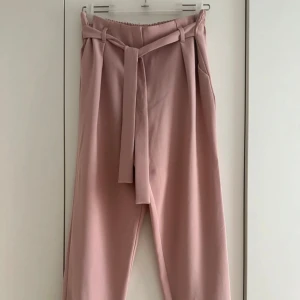 Puderrosa byxor med knytband - Snygga puderrosa byxor med avtagbart knytband i midjan. Byxorna har raka ben, resår baktill. Perfekta för en chill men ändå uppklädd look.  Byxorna är i storlek 10