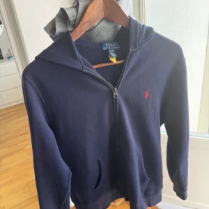 Marinblå hoodie från Polo Ralph Lauren - Snygg marinblå hoodie från Polo Ralph Lauren med dragkedja och klassisk röd logga på bröstet. Tröjan har huva, fickor framtill och är gjord i mjuk bomullsblandning. Perfekt för en chill och stilren look. Använd bara fåtal gånger, köpte koftan för 1299 från KidsBrandStore ✅🤝