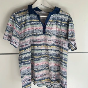 Färgglad randig t-shirt med krage - Säljer en unik t-shirt med färgglada ränder i blått, gult, rosa och vitt. T-shirten har korta ärmar, blå krage och knappslå med tre knappar framtill. Perfekt för dig som gillar mönster och vill sticka ut lite extra.