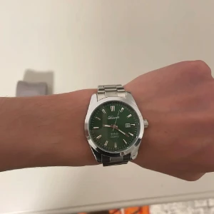 Grön herrklocka med stållänk från Doventi  - Stilren herrklocka från Doventi med grön urtavla, silverfärgade index och datumvisning. Klockan har en robust länk i metall och klassisk rund boett. Perfekt accessoar för dig som gillar en modern och clean look.