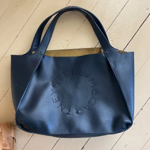  Stella McCartney logo bag - Stilren svart shoppingväska från Stella McCartney i syntetmaterial med perforerad logga framtill. Väskan har två rejäla handtag och rymligt fack med beige insida samt en avtagbar innerficka. materialet är lite slitet på vissa ställen