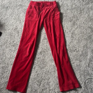 Röda mjukisbyxor Juicy Couture - Snygga röda mjukisbyxor från Juicy Couture i mjuk velour med broderad JC-logga på fickan. Byxorna har snörning i midjan, stora fickor fram och bakfickor med knappar. Perfekta för en chill och trendig look.